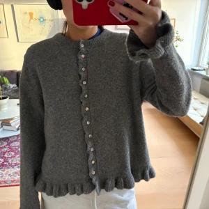 Grå kofta med volang från Zara - Superfin grå kofta från Zara med volangkant vid ärmslut och nederkant. Koftan har pärlemorknappar framtill och är stickad i mjukt material. Perfekt för lager-på-lager och ger en söt touch till din outfit. Pris kan diskuteras 