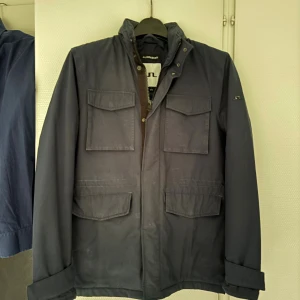 Mörkblå parkajacka från J.Lindeberg - Parkajacka från J.Lindeberg i mörkblått med fyra stora fickor framtill och hög krage. Jackan har dragkedja och tryckknappar, samt diskret JL-logga på ärmen. Perfekt för dig som gillar stilren och funktionell design.