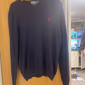 Mörkblå stickad tröja Polo Ralph Lauren - Mörkblå stickad tröja från Polo Ralph Lauren i slim fit-modell. Klassisk design med rund halsringning och rött broderat logotyp på bröstet. Perfekt för dig som gillar stilrena och tidlösa plagg. Matrialet är Merino 