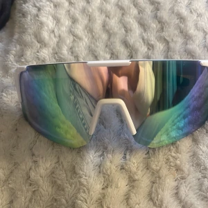 Vita sportiga solglasögon med spegelglas - Säljer ett par feta sportglasögon med vit båge och stora spegelglas i grönt och gult. Glasögonen har en futuristisk, kantig form och svarta detaljer på insidan. Perfekta för dig som vill sticka ut när du tränar eller cyklar.
