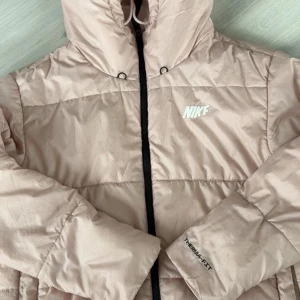 Rosa pufferjacka från Nike Therma-Fit - Säljer en Rosa pufferjacka från Nike med Therma-Fit-teknologi. Jackan har huva, svart dragkedja och Nike-logga på bröstet. Perfekt för kyliga dagar och riktigt skön att bära. Klassisk och stilren design som funkar till det mesta.