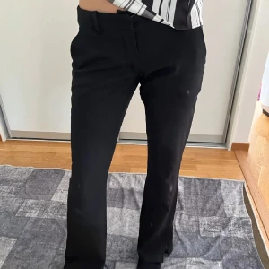 Svarta kostymbyxor med bootcut - Snygga svarta kostymbyxor med klassisk bootcut och hög midja. Byxorna har diskreta fickor bak och bälteshällor i midjan. Perfekta för en clean och stilren look. Materialet känns mjukt och följsamt, vilket gör dem bekväma hela dagen.