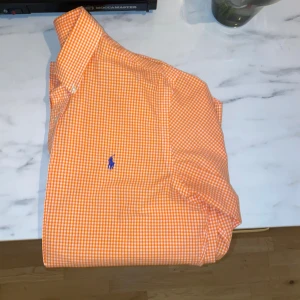 Orange rutig skjorta Polo Ralph Lauren - Orange och vit rutig skjorta från Polo Ralph Lauren i slim fit. Skjortan har klassisk krage, långa ärmar och knäppning framtill. Broderad blå logga på bröstet. Tillverkad i mjuk bomull, perfekt för en fräsch och stilren look.