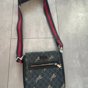 Goc c i axelväska med hundmönster - Snygg axelväska från pucci med klassiskt GG-mönster och små hundar tryckta över hela väskan. Väskan har en svart bas, dragkedja framtill och bred axelrem i rött och marinblått. Perfekt för dig som vill sticka ut med en unik designeraccessoar.