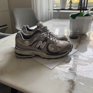 New Balance 2002R camo sneakers - Säljer ett par New Balance 2002R sneakers i grått och beige camo-mönster. Skorna har klassisk N-logga på sidan, snörning och en chunky sula med coola detaljer. Ovandelen är i en mix av mocka och syntetmaterial. Perfekta för dig som gillar streetwear och vill sticka ut.