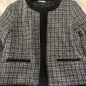 Rutig kavaj från Vila, svartvit, stl 38 - Snygg svartvit rutig kavaj från Vila i storlek 38. Jackan har klassisk krage, raka fickor med svarta detaljer och är tillverkad i ett strukturerat tyg som ger en exklusiv känsla. Perfekt för dig som vill ha en stilren och trendig look.