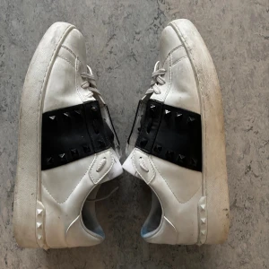 Valentino Rockstud sneakers vit/svart - Säljer ett par Valentino Rockstud sneakers i vitt skinn med bred svart rem över vristen dekorerad med svarta nitar. Klassisk rund tå, platt sula och snörning. Snygg kontrast mellan vitt och svart, samt coola detaljer på sidor och häl. nån ska använda och i bra skick