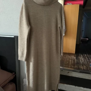 Beige stickad poloklänning - Mysig beige stickad långklänning med polokrage och långa ärmar. Klänningen har ribbade muddar och en liten slits i nederkanten på ena sidan. Perfekt för kyliga dagar och enkel att styla med boots eller sneakers.