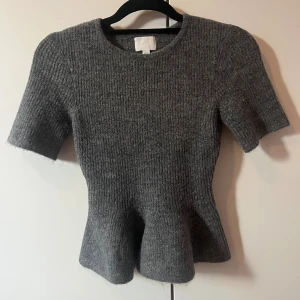 Stickad topp från h&m💓💓 - Helt ny och superfin grå stickad topp från H&M i storlek XS. Toppen har kort ärm, rund halsringning och markerad midja med lätt utsvängd peplumkant. Prislappen är kvar och tröjan har aldrig använts 🩷