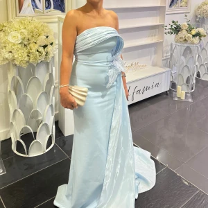 Ljusblå axelbandslös långklänning - Elegant ljusblå axelbandslös långklänning med draperad överdel och vacker spetsdetalj i midjan. Klänningen har en figurnära passform och ett svepande tyg som ger en lyxig känsla. Perfekt för bal eller speciella tillfällen.