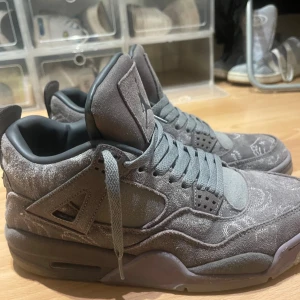 Nike Air Jordan 4 KAWS grå mocka - Säljer ett par Nike Air Jordan 4 KAWS i grå mocka med unika broderade detaljer och ikoniska XX på hälen. Skorna har snörning, platt sula och rund tå. Perfekta för dig som vill sticka ut med en limiterad sneaker i snygg mörkgrå färgton.