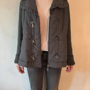 Grå quiltad jacka med stickade ärmar - Snygg grå jacka med quiltad front och rygg samt stickade ärmar. Jackan har dragkedja och coola metallhakar framtill. Passformen är normal och ärmarna är långa och ribbstickade.