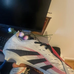Adidas vita och svarta fotbollsskor - Säljer ett par Adidas fotbollsskor i vitt med svarta och röda detaljer. Skorna har klassisk snörning, låg profil och mönstrad sula för bra grepp på planen. Tungan är röd med Adidas-logga och ovandelen är i syntetmaterial. Perfekta för dig som vill sticka ut på fotbollsplanen. Ena tungan är sönder som ni ser darför så låg pris 