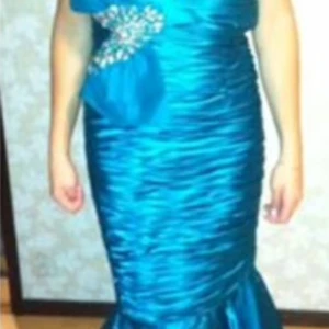 Turkos långklänning med strassdetalj - Säljer en turkos långklänning i glansigt material med en axel och snygga strassdetaljer vid axeln. Klänningen har figurnära passform och volang nertill för extra wow-känsla. Perfekt för dig som vill sticka ut med färg och glamour.