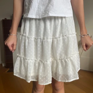 Vit volangkjol från FB Sister XS - Supersöt vit kortkjol från FB Sister i storlek XS. Kjolen har flera volanger och ett diskret prickigt mönster i tyget. Den är lätt och luftig, perfekt för varma dagar. Resår i midjan för extra komfort och snygg passform. 