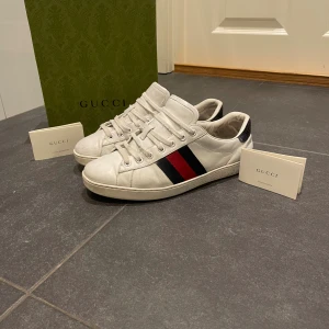 Vita Gucci ace med blå röda detaljer - Tja säljer nu mina Gucci ace sneakers  i vitt skinn med klassiska röd och blå ränder på sidan. Baksidan har smärkblå  ormskinnsmönstrad detalj och logga. Skorna har snörning och platt sula, perfekt för en clean och lyxig look.