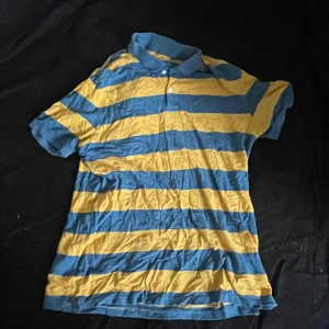 Randig blå och gul pikétröja - Säljer en kortärmad pikétröja med breda blå och gula ränder. Tröjan har klassisk krage och knappar framtill. Perfekt för dig som gillar färg och vill sticka ut lite extra. Skön och luftig modell som passar till många olika stilar.