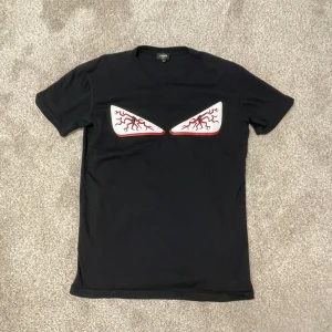 Fendi T-shirt - Svart t-shirt från Fendi med korta ärmar och ett unikt ögonmotiv framtill i vitt och rött. Tillverkad i mjuk bomull med lite elastan för extra komfort. T-shirten är i väldigt bra skick. Storleken är L men passar M bra (pris kan diskuteras).