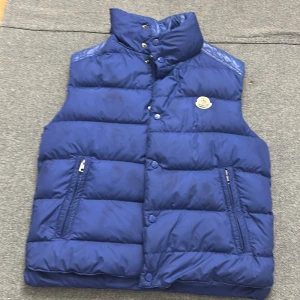 Blå dunväst från Moncler - Snygg blå dunväst från Moncler med hög krage och två dragkedjefickor framtill. Västen har quiltad design och klassisk Moncler-logga på bröstet. Perfekt för lager-på-lager och ger en sportig vibe.