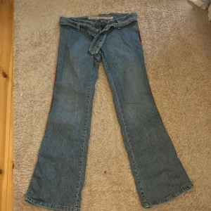 Blå bootcut jeans Mustang Superior - Snygga blå jeans från Mustang Superior Denim med bootcut passform och klassisk jeanslook. Byxorna har ett tillhörande jeansbälte med metallspänne och röda detaljer längs sidorna. Perfekta för dig som gillar retrovibbar och vill sticka ut lite extra. Säljer då d inte komemr till användning.