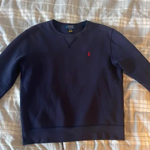 Mörkblå sweatshirt från Polo Ralph Lauren - Mörkblå sweatshirt från Polo Ralph Lauren med klassisk rund hals och rött broderat logotyp på bröstet. Tröjan har lång ärm och är tillverkad i mjukt bomullsmaterial. Perfekt för en chill och stilren look. ❗️Pris kan diskuteras❗️