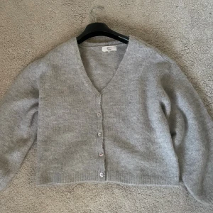 Grå kofta från Gina Tricot - Mysig grå kofta från Gina Tricot med v-ringning och knappar framtill. Koftan är stickad, har en loose passform och långa ärmar. Perfekt att slänga över en topp för en chill look. Superenkel att styla och passar till allt.