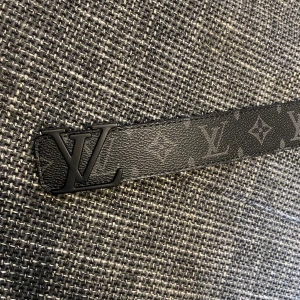 Svart Louis Vuitton monogram bälte - Snyggt svart bälte från Louis Vuitton med klassiskt monogrammönster och mattsvart LV-spänne. Tillverkat i exklusivt skinn med tydlig struktur och diskreta grå detaljer i mönstret. Perfekt accessoar för att lyfta din outfit med en lyxig touch.