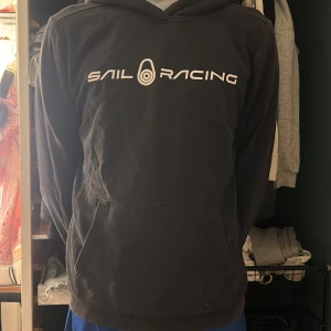 Svart hoodie från Sail Racing - Svart hoodie från Sail Racing med vit logga framtill. Klassisk modell med huva och känguruficka. Mjuk och skön, perfekt för chill dagar. Långärmad och i bomullsmix som känns bekväm mot huden.