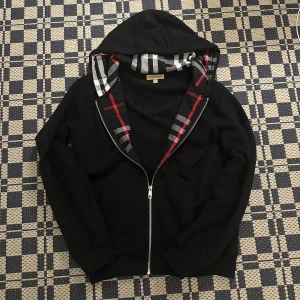 Svart Burberry hoodie med dragkedja - Svart hoodie från Burberry med klassiskt rutigt foder i huvan och silverfärgad dragkedja. Tröjan har snörning i huvan, två fickor fram och diskret broderad logga på bröstet. Perfekt för dig som gillar stilren streetwear med lyxig touch.