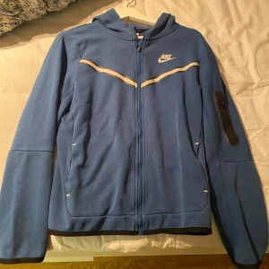 Blå hoodiejacka från Nike  - Nike hoodiejacka i blått med vit logga och vita detaljer över bröstet. Jackan har dragkedja hela vägen, huva och en praktisk ficka med dragkedja på ena ärmen. 