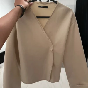 Beige omlottkofta från Gina Tricot - Säljer en beige omlottkofta från Gina Tricot i storlek XS. Koftan har en snygg v-ringning, markerade sömmar längs kanterna och är tillverkad i ett mjukt material. Perfekt för lager-på-lager och enkel att styla till olika outfits.