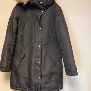 Svart parkajacka med fuskpäls - Svart parkajacka med huva och fuskpälsdetalj. Jackan har dragkedja och knappar framtill, två snedställda bröstfickor med dragkedja och två stora fickor med lock. Perfekt för kalla dagar och riktigt snygg stil.