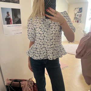Mönstrad blus med volang från Lindex - Supersöt blus i vitt med blått blommigt mönster från Holly & Whyte by Lindex. Blusen har puffiga korta ärmar, volang nedtill och knytning vid halsen. Perfekt för dig som gillar romantisk stil och detaljer. Materialet är mjukt och lätt bomull.