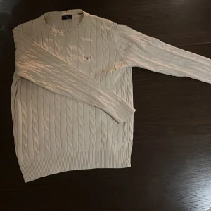 Vit kabelstickad tröja från Gant - Säljer en klassisk vit kabelstickad tröja från Gant med rund halsringning och diskret logga på bröstet. Tröjan har lång ärm och ribbade muddar vid ärmslut och nederkant. Perfekt för en clean och stilren look.strl 164 13-14 år 