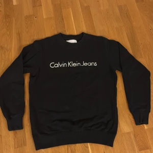 Svart sweatshirt från Calvin Klein Jeans - Svart sweatshirt från Calvin Klein Jeans med vit logga framtill. Klassisk rund halsringning och ribbade muddar vid ärmslut och nederkant. Tillverkad i mjuk bomullsmix som är skön att ha på sig. Perfekt för en clean look.