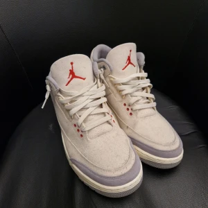 Nike Air Jordan 3 muslin - Säljer ett par Nike Air Jordan 3 sneakers i beige canvas med lila detaljer och röd Jumpman-logga. Skorna har snörning, rund tå och platt sula. Perfekta för dig som gillar klassiska och stilrena sneakers med en twist.