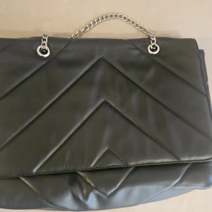 Svart quiltad axelväska från NLY - Snygg svart axelväska från NLY Accessories med quiltad design och silverfärgad kedja som axelrem. Väskan har ett stilrent mönster med diagonala sömmar och är gjord i skinnimitation. Perfekt storlek för att få plats med allt viktigt.