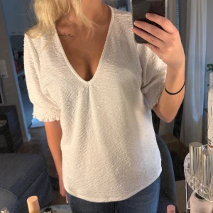 Vit blus med v-ringning från Vero Moda - Säljer en vit blus från Vero Moda i storlek XL. Blusen har en djup v-ringning, korta ärmar med smockdetalj och ett lätt strukturerat tyg. Passformen är normal och materialet känns mjukt och luftigt, perfekt för vår och sommar.