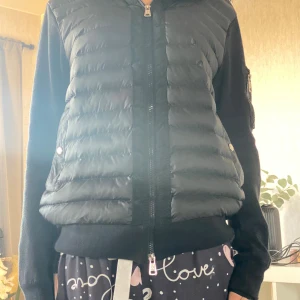 Svart Moncler pufferjacka med stickade ärmar - Snygg svart pufferjacka från Moncler med quiltad framsida och stickade ärmar. Jackan har dragkedja framtill, fickor med tryckknappar och en extra ficka med dragkedja på ärmen. Perfekt för dig som vill ha en stilren och exklusiv look.