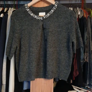 Grå kofta med pärldetaljer - Mysig grå kofta i stickad ullmix med kort ärm och knappar framtill. Runt halsringningen sitter glittrande pärldetaljer som ger en extra touch. Perfekt för lager-på-lager och när du vill sticka ut lite extra.