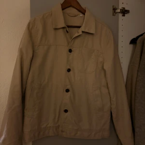 Beige overshirt med knappar - Snygg beige overshirt med klassisk krage och knäppning framtill. Jackan har tre fickor, varav en på bröstet, och är tillverkad i ett mjukt bomullsmaterial. Perfekt för lager-på-lager och enkel att styla till olika outfits.