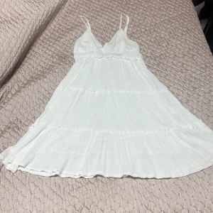 Vit somrig kortklänning med volang - Luftig vit kortklänning med tunna axelband och v-ringad hals. Klänningen har volangdetaljer och är tillverkad i ett lätt, plisserat tyg som ger en flowy känsla. Perfekt för varma dagar och en avslappnad stil.