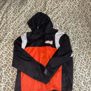 Jordan vindjacka - Snygg vindjacka från Jordan i svart, rött och vitt med huva och stora loggor både fram och bak. Jackan har långa ärmar, elastiska muddar och är tillverkad i lätt polyester. Perfekt för dig som gillar sportig streetstyle. Storlek XXL