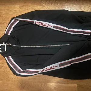 Svart track jacket från Gucci - Svart track jacket från Gucci med vita och röda ränder längs ärmarna och tydlig logga. Jackan har hel dragkedja framtill, hög krage och ribbade muddar. Perfekt för en sportig och trendig look.
