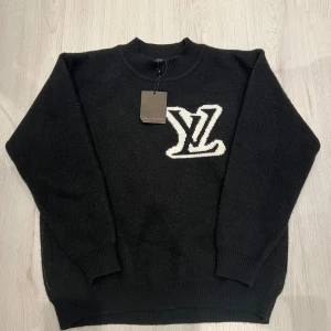 Svart stickad tröja Louis Vuitton - Svart stickad tröja från Louis Vuitton med stort vitt LV-logo både fram och bak. Tröjan har rund halsringning, ribbade muddar och en klassisk passform. Perfekt för dig som vill ha en lyxig och stilren look med tydlig designer-touch.