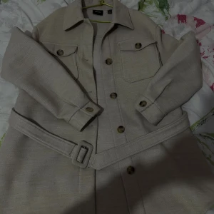 Beige skjortklänning från Vero Moda - Stilren beige skjortklänning från Vero Moda med klassisk krage, knappar framtill, två bröstfickor och matchande bälte i midjan. Långa ärmar med knappdetalj vid manschetten. Perfekt för dig som gillar minimalistisk och clean stil.