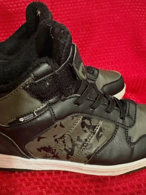 Svarta och gröna sneakers från Warp - Säljer ett par svarta och mörkgröna sneakers från Warp i storlek 34. Skorna har högt skaft, snörning och kardborreband upptill. Sidorna har coolt camouflage-mönster och insidan är fodrad med mjukt syntetmaterial. Perfekt för dig som gillar streetstyle.