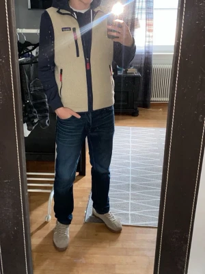 Beige fleeceväst från Patagonia - Säljer en beige fleeceväst från Patagonia med marinblå detaljer och röda dragkedjor. Västen har tre fickor med dragkedja och en hög krage. Perfekt att slänga på över en hoodie för extra värme. Klassisk och stilren design.