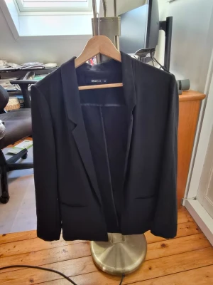 Svart kavaj från Gina Tricot - Stilren svart kavaj från Gina Tricot i storlek 34. Klassisk modell med smala slag, två fickor framtill och långärmad design. Perfekt för dig som vill ha en enkel och snygg look. Materialet känns mjukt och har en lätt struktur.