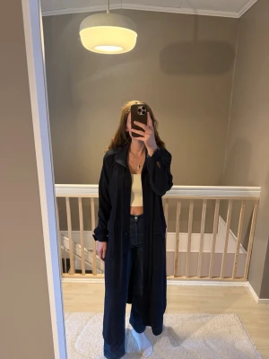 Mörkblå trenchcoat från Rodebjer - Säljer en lång och stilren mörkblå trenchcoat från Rodebjer. Jackan har knappar hela vägen, bälte i midjan och klassisk krage. Materialet är mjukt och känns som bomull, perfekt för vår och höst. Långärmad och med snygg passform som ger en cool vibe.